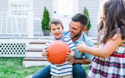 Laughing boy holding an orange ball with his father and sister in front of their house