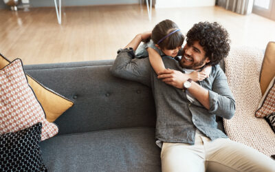 Smiling man on a gray couch hugging a young girl who has her arms around his neck